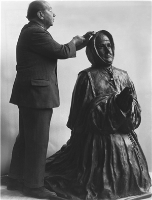 A man in a suit with his hand on the head of a stone sculpture of a kneeling, praying nun